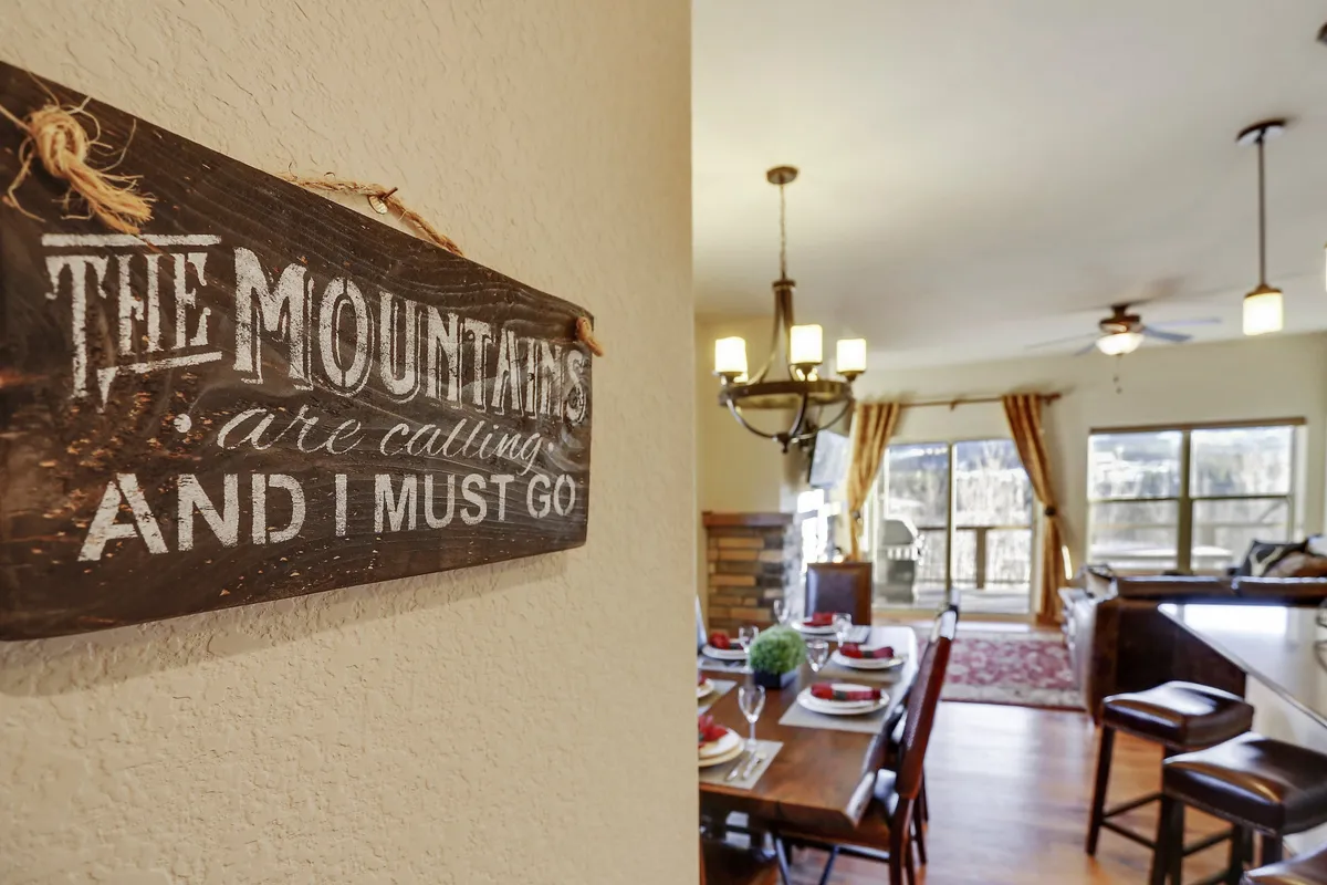 A cozy dining area featuring a wooden sign, a dining table with place settings, and large windows revealing scenic views. The atmosphere blends comfort, gathering, and a sense of connection to the outdoors, appealing in all seasons.
