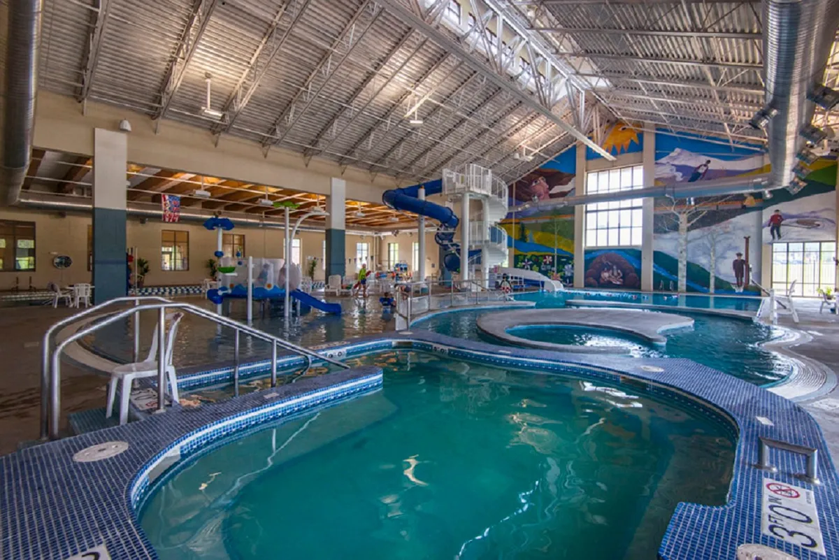 A lively indoor recreation center with a water slide, pool areas, a colorful mural, and children playing, capturing the energy of family-friendly fun and relaxation.
