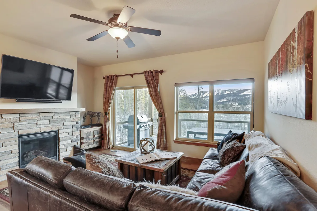 This image features a large leather sectional sofa positioned in front of a striking stone fireplace, with expansive windows that frame picturesque mountain views. Decorative wall art adds a touch of personality, while a coffee table with decorative items is set centrally, evoking a cozy and inviting environment ideal for gatherings, relaxation, and enjoying scenic sunsets.