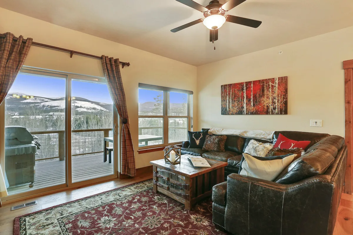 The image showcases a spacious, inviting living room with a comfortable sofa and coffee table arranged before expansive windows framing breathtaking mountain views. Warm natural light pours in, creating a cozy atmosphere perfect for relaxing, hosting friends, or unwinding with a book. Tasteful artwork and a fireplace add to the charm, suggesting evenings spent watching sunsets and sharing stories by the fire.