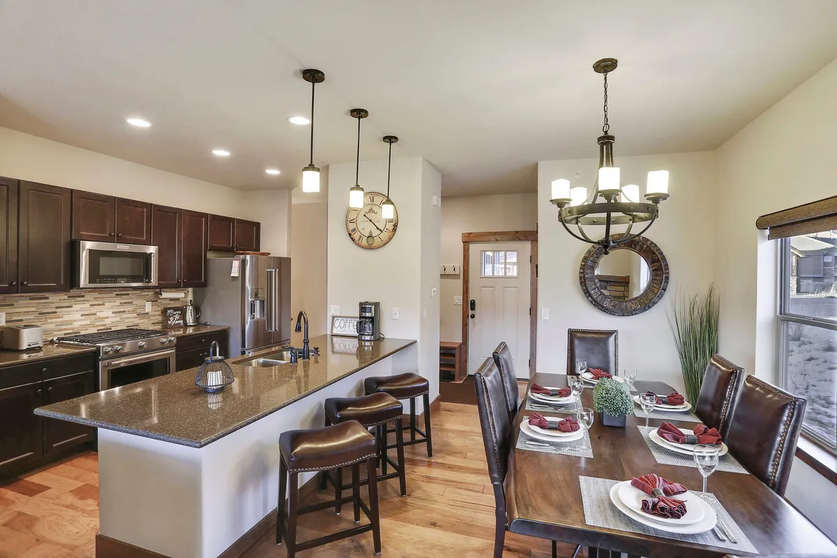 This scene focuses on the kitchen island and dining table, illuminated by a stylish chandelier and accented by a large clock on the wall. The setup invites communal cooking, social dining, and festive celebrations, further illustrating the home's role as a hub for shared experiences and memory-making.