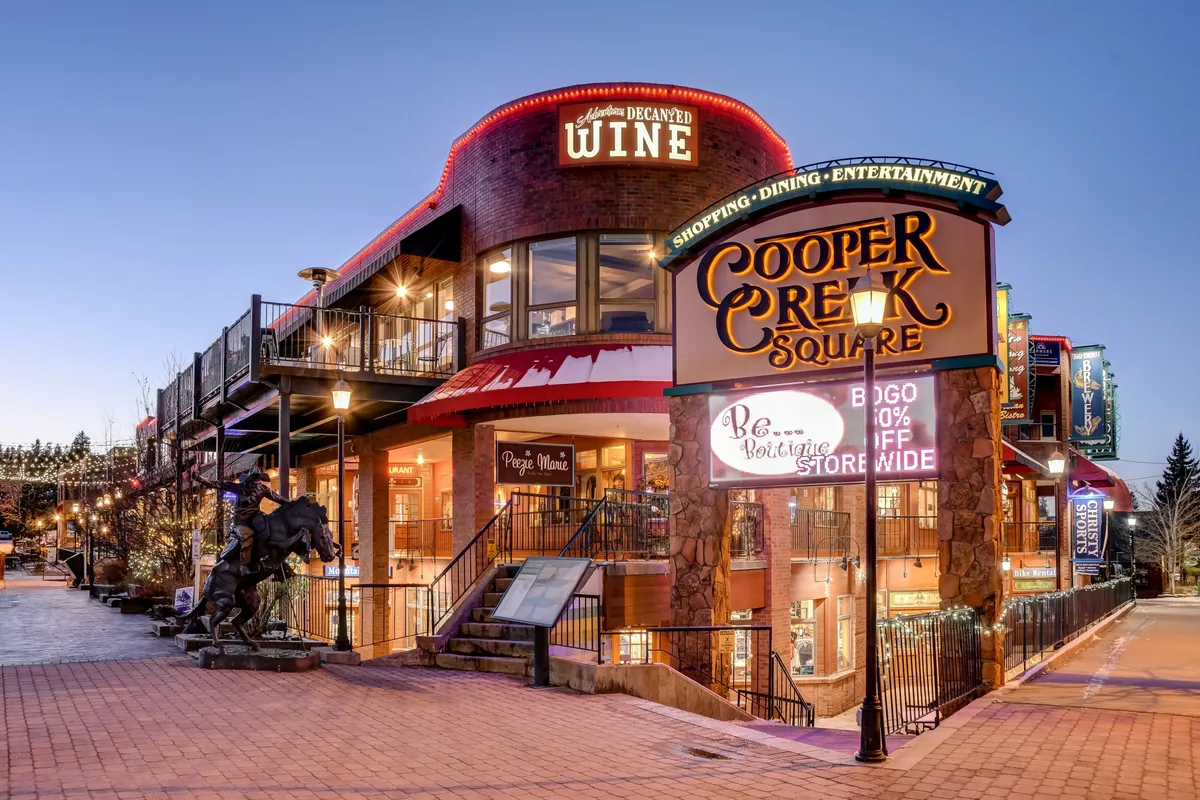 A bustling exterior scene of Cooper Creek Square, highlighted by outdoor seating, a prominent sign, a wine store, and a sculptural horse. The area appears lively and welcoming, with the promise of live music, art displays, and social interaction.