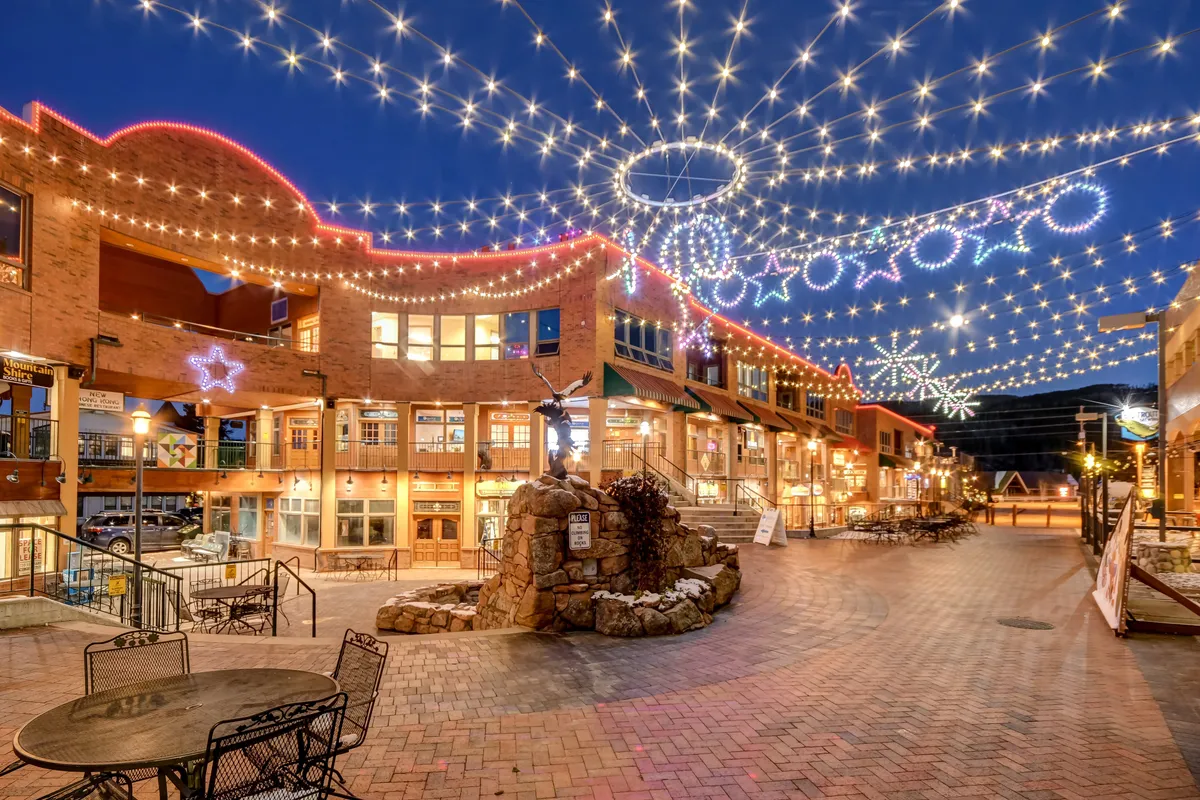A lively, colorful outdoor plaza featuring decorative string lights, a stone fountain, and inviting outdoor seating. The facades of nearby restaurants and shops add a festive ambiance, making it clear this is a hub for social gatherings and community events.